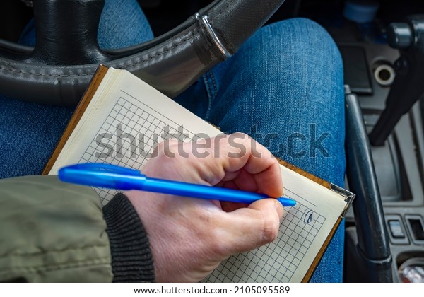 an open paper notebook, a book for\
notes, a journal for notes lies on the driver lap inside the car,\
on which a man, a male, makes notes with a ballpoint\
pen