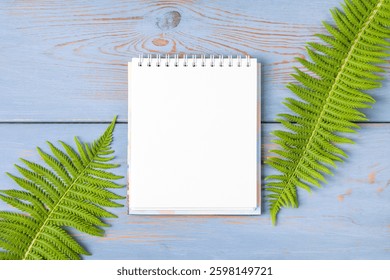 Open paper notebook with empty page with green fern leaves on a wooden blue table. Top view, flat lay, work space on blue background. Hello spring