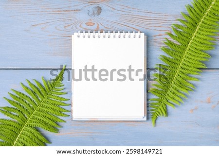 Open paper notebook with empty page with green fern leaves on a wooden blue table. Top view, flat lay, work space on blue background. Hello spring