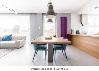 Open Plan Apartment Of Family Living-space With Wooden Kitchen Countertop, Purple Radiator, Communal Table With Turquoise Chairs And Spacious Living Room With Beige Couch