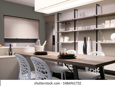 Open Plan Kitchen And Dining Room With Neutral Beige Decor, Shelving Wall Unit And A Modern Table And Chairs Lit By A Large Overhead Ceiling Light