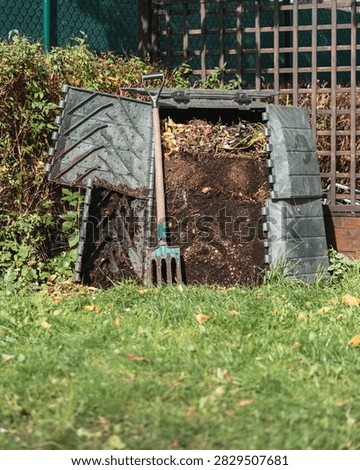 An open plastic compost bin in a backyard garden with a garden fork leaning against it, showing layers of decomposing kitchen and garden waste on top of dark, rich compost soil, ready for turning.