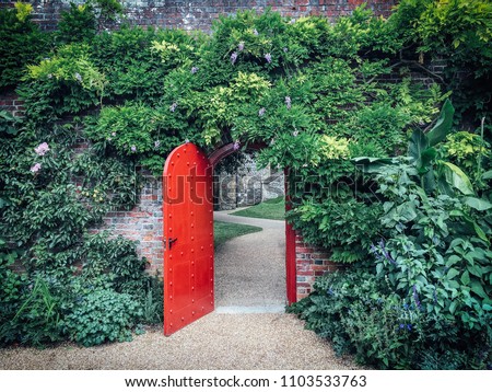 Open, red, half-round door in a brick wall covered with green vines, behind a door a winding gravel path. Lush green foliage cover the brick wall. A mysterious path leads towards the castle walls.