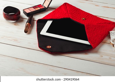 An Open Red Lady's Bag With Tablet Computer In A White Wooden Table.
Near The Handbags Are Women's Accessories. Mascara, Eye Shadow, Watch And Bracelet