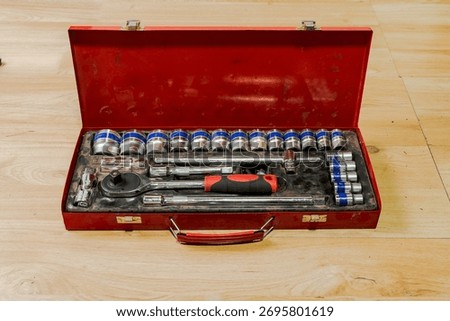 Open red metal toolbox with socket wrench set on wooden floor, featuring ratchet handle, socket extensions, and T-bar in organized mechanical repair setup.