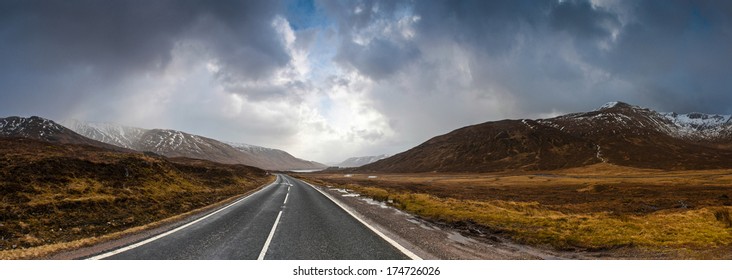 Open Road Leading Through The Scottish Highlands Of Glen Coe, Snowcapped Mountains And Dramatic Storm Broken Sky.