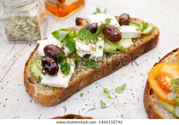 Open sandwich made of\
slices of sourdough bread with avocado, feta cheese, kalamata\
olives, olive oil and oregano on a wooden white table, close-up.\
Vegetarian food