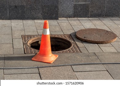 Open sewer manhole cover and traffic cone on a city street. Sewerage repair. Emergency service and accident. Drainage system maintenance. Danger sign