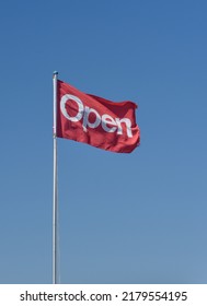 Open Sign, Red Flag With White Writing Against A Plain Blue Sky.  