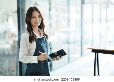 open a small business Portrait of happy Asian woman in apron holding tablet in front of cafe - Shutterstock ID 2258461389