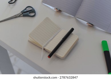 Open small lined notebook with black pen, scissors, green highlighter on white surface, larger notebook in background