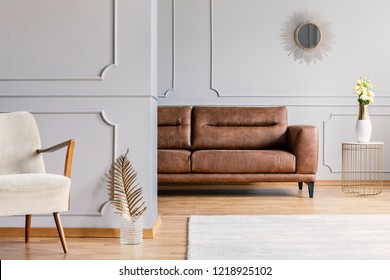 Open space living room interior with decorative mirror on wall with wainscoting, brown leather sofa, fresh roses on end table and gold fern leaf in glass vase placed by white armchair in real photo