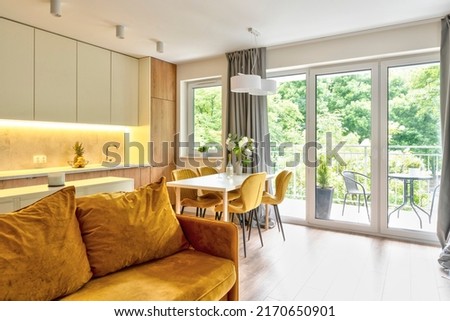 Open space living room with kitchen and big balcony window, mustard comfortable sofa with pillow and kitchen furniture. Wooden floor in modern apartment. Architecture of indoors. [[stock_photo]] © 