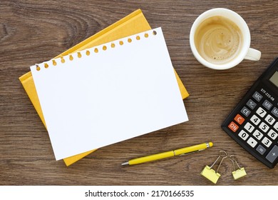 Open Spiral Blank Notebook With Calculator, A Yellow Pen, A Cup Of Coffee And A Small Flower Plant On A White Desk, Flat View From Above