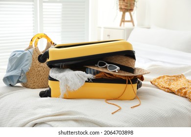 Open Suitcase Full Of Clothes And Summer Accessories On Bed In Room