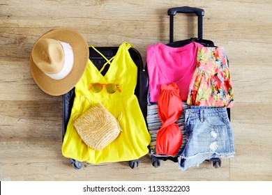 Open suitcase fully packed with folded women's clothing and accessories on the floor. Woman packing for tropical vacation concept. Female luggage w/ things. Background, close up, copy space, top view.