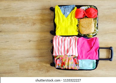 Open Suitcase Fully Packed With Folded Women's Clothing And Accessories On The Floor. Woman Packing For Tropical Vacation Concept. Female Luggage W/ Things. Background, Close Up, Copy Space, Top View.