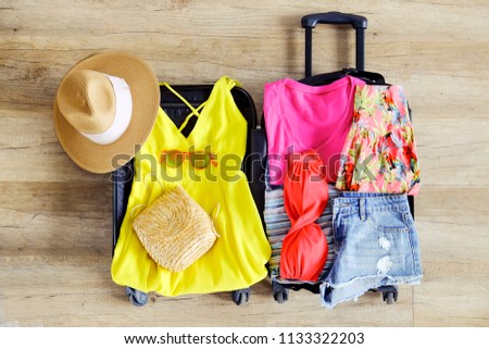 Open suitcase fully packed with folded women's clothing and accessories on the floor. Woman packing for tropical vacation concept. Female luggage w/ things. Background, close up, copy space, top view.