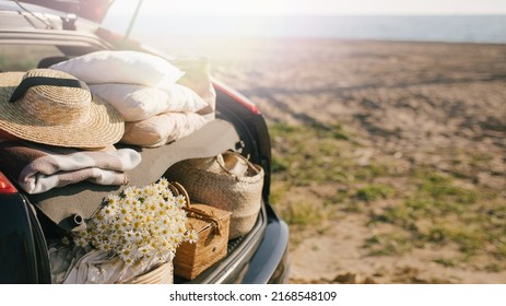 Open Trunk Of Car With Picnic Items: Straw Hat, Basket With Food And Flowers With Summer Beach On The Background. Place For Text.