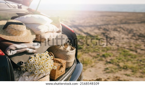 Open trunk of car with picnic items: straw hat,\
basket with food and flowers with summer beach on the background.\
Place for text.
