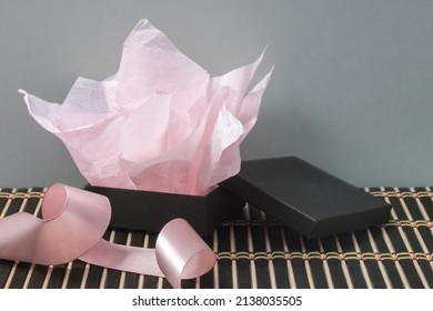 An open, unwrapped black jewelry gift box with pink tissue paper and satin ribbon, on a striped bamboo mat with a gray background.
