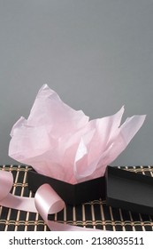 An Open, Unwrapped Black Jewelry Gift Box With Pink Tissue Paper And Satin Ribbon, On A Striped Bamboo Mat With A Gray Background. Vertical Image With Copy Space Above.