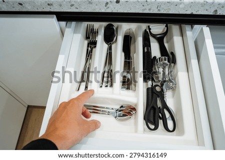 Open white kitchen drawer filled with stainless steel forks, spoons, knives and a section of serving tongs. adjacent compartment holds blackhandled kitchen