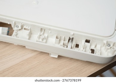 Open white printer and scanner casing interior plastic compartments, spring mechanism and ribbon cable on a wooden desk. Electronics repair, device maintenance, teardown and technical illustration.