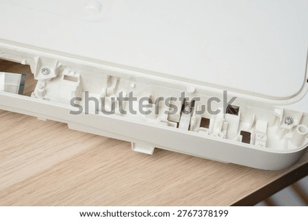 Open white printer and scanner casing interior plastic compartments, spring mechanism and ribbon cable on a wooden desk. Electronics repair, device maintenance, teardown and technical illustration.