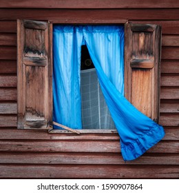 Open Window Of A Brown Wooden House. A Blue Curtain Is Hanging In The Window, Partly Outside Blowing In The Wind. Wooden Window Shutters. No Glass. Mosquito Net In The Room