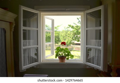 An Open Window With A Geranium Flower  Inside An Old Vintage Village Rural Wooden Cottage House, With A View Of A Green Countryside Outside 