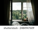 An open window with a view of a green forest through white curtains. A bed with gray linens is in the foreground, partially out of focus. A potted plant and other objects are on the windowsill. 