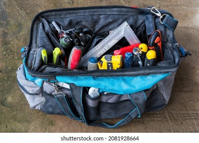 An Open Work Bag With A Variety Of Repair Tools Sits On The Concrete Floor. The Surface Of The Used Items Is Covered With Construction Dust. Background. 