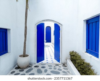 An opened blue door with white wall, closed windows, and green bushes. Santorini Greece architecture style. - Powered by Shutterstock