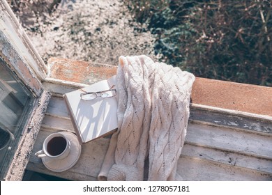 Opened Book, Cup Of Tea Or Coffee, Glasses And Knitted Sweater On Window Sill Over Blooming Tree View. Cozy Spring Weekend Concept. The Text On Pages Is Not Recognizable.