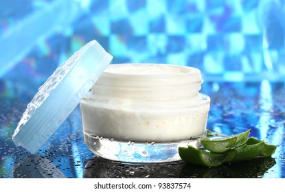 Opened glass jar of cream and aloe on blue background with water droplets