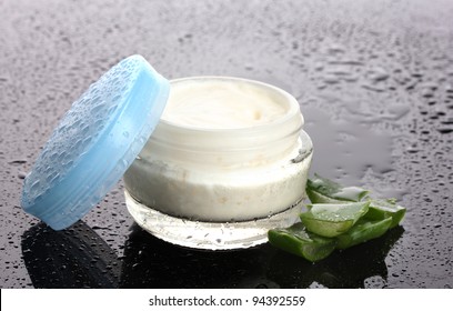 Opened glass jar of cream and aloe on black background with water droplets