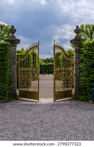 Opened golden gates with intricate scrollwork stand between stone pillars, leading to an open gravel path within Herrenhausen Gardens.