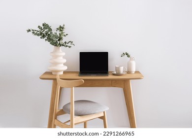 opened laptop, ceramic vase with eucalyptus branch and cup of coffee on wooden desk near comfortable chair on white wall background in stylish apartment - Powered by Shutterstock