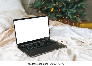 Opened laptop with white screen on bed in bedroom with decorated Christmas tree at home, mockup 