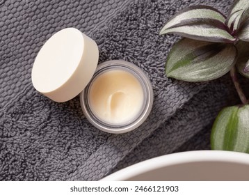 Opened lip balm jar with blank lid on dark grey folded towels, near green plant and vessel basin in bathroom, top view, cosmetic brand packaging mockup. Men facial cream
 - Powered by Shutterstock