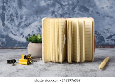 Opened notebook, flower in pot, binder clips and pen on grunge table near grey wall