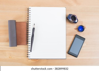 Opened notebook or notepad with pen, mobile and ink on wooden desk.