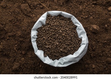 Opened plastic bag with black granules of chicken manure on dark soil background. Closeup. Product for root feeding of vegetables, flowers and plants. 