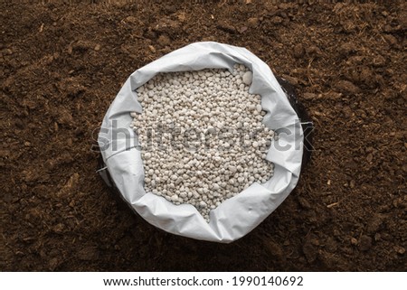 Opened plastic bag with gray complex fertiliser granules on dark soil background. Closeup. Product for root feeding of vegetables, flowers and plants. 