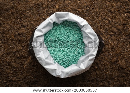 Opened plastic bag with green complex fertiliser granules on dark soil background. Closeup. Product for root feeding of vegetables, flowers and plants. 