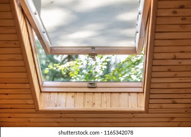 Opened Roof Window With Blinds Or Curtain In Wooden House Attic. Room With Slanted Ceiling Made Of Natural Eco Materials And Park View Through Opened Window. Environment Friendly House