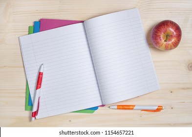 Opened School Notebook With Pens And Apple On The Wooden Desk. Top View With Empty Place For Text