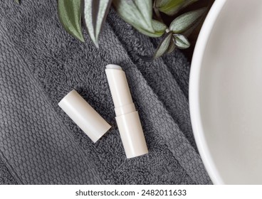 Opened white blank lip balm stick on dark grey folded towels near green plant and vessel basin, top view, cosmetic mockup.  Lifestile scene with skincare beauty product - Powered by Shutterstock