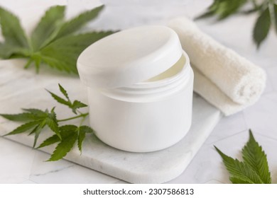 Opened white cream jar with a lid near green cannabis sativa leaves on a marble table. Cosmetic Mockup, Copy space. Organic skincare beauty product. Eco friendly body or hand cream with hemp